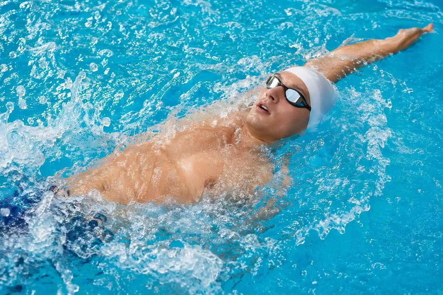 Junger Schwimmer mit Schwimmkappe und Schwimmbrille schwimmt Rückenschwimmen in einem klaren, blauen Schwimmbecken