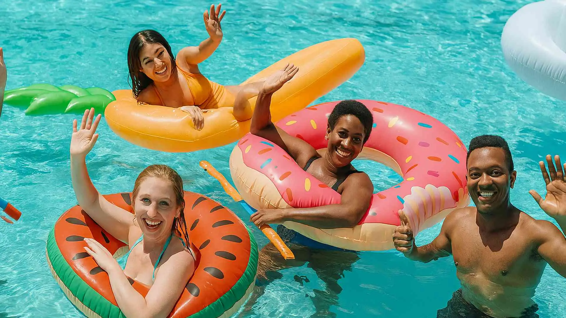 Mehrere fröhliche Menschen in einem Pool mit bunten Schwimmringen, sie winken und genießen das Wasser